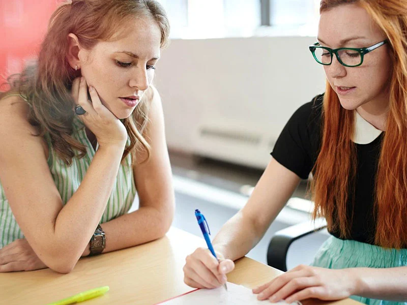 Two women sitting at a table writing on paper.