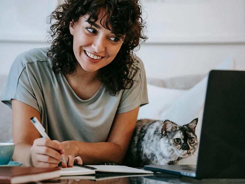 A woman smiles while writing on paper next to her laptop.